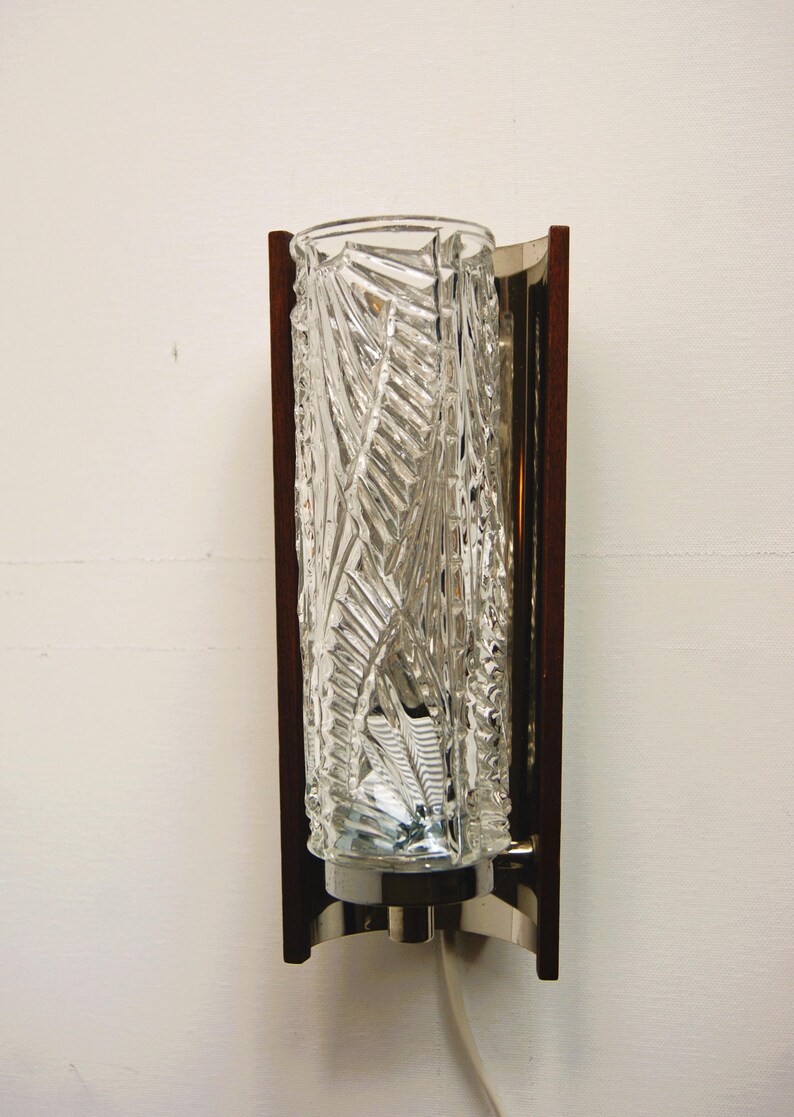 K&ouml;nnte beinhalten: Eine Wandleuchte im Stil der Mid-Century Modern mit einem klaren Glasschirm mit geometrischem Muster. Die Leuchte hat einen Rahmen aus Holz und Metall.