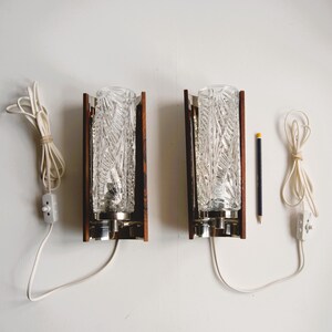 K&ouml;nnte beinhalten: Zwei vintage Wandleuchten mit klaren Glasschirmen und Holzrahmen. Jede Leuchte hat ein wei&szlig;es Kabel mit einem Schalter.
