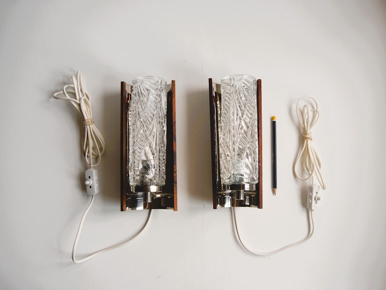 K&ouml;nnte beinhalten: Zwei vintage Wandleuchten mit klaren Glasschirmen und Holzrahmen. Jede Leuchte hat ein wei&szlig;es Kabel mit einem Schalter.