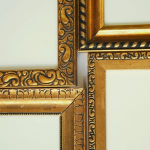 May include: Close-up of four ornate gold picture frames with white interiors. The frames feature intricate, dark-toned carvings and a glossy finish. The corners of the frames are visible, showcasing the detailed craftsmanship.