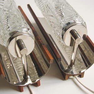 K&ouml;nnte beinhalten: Zwei silberne Wandleuchten mit Glasschirmen. Die Schirme haben ein strukturiertes, blattartiges Muster. Die Leuchten sind auf Holzsockeln montiert.