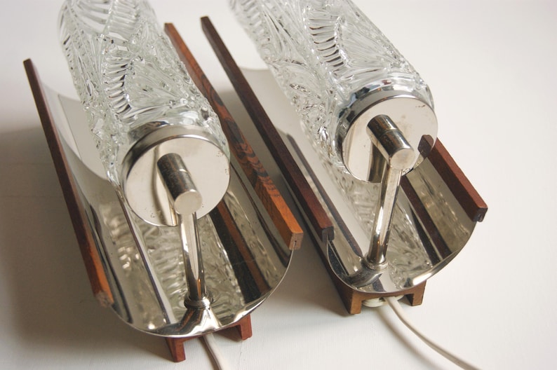 K&ouml;nnte beinhalten: Zwei silberne Wandleuchten mit Glasschirmen. Die Schirme haben ein strukturiertes, blattartiges Muster. Die Leuchten sind auf Holzsockeln montiert.