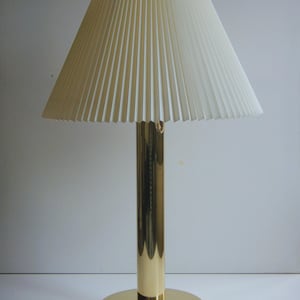 May include: A gold-colored table lamp with a pleated off-white lampshade. The lamp has a cylindrical base and a round base. The lampshade is cone-shaped and made of vertical pleats. The lamp is designed for indoor use.