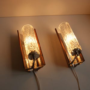 K&ouml;nnte beinhalten: Paar Wandleuchten im Mid-Century-Modern-Stil mit Holz- und Chrom-Akzenten. Jede Leuchte verf&uuml;gt &uuml;ber einen klaren Glasschirm mit geometrischem Muster.