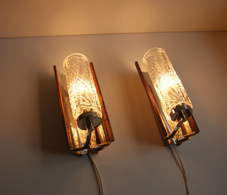 K&ouml;nnte beinhalten: Paar Wandleuchten im Mid-Century-Modern-Stil mit Holz- und Chrom-Akzenten. Jede Leuchte verf&uuml;gt &uuml;ber einen klaren Glasschirm mit geometrischem Muster.
