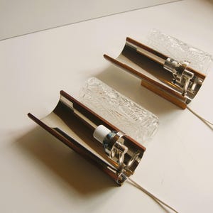 K&ouml;nnte beinhalten: Zwei Wandleuchten aus Chrom und Holz mit klaren, strukturierten Glasschirmen. Die Leuchten haben ein geschwungenes Design und sind mit wei&szlig;en Kabeln verdrahtet.