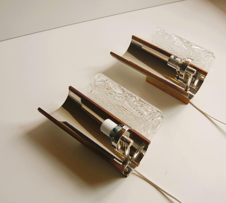 K&ouml;nnte beinhalten: Zwei Wandleuchten aus Chrom und Holz mit klaren, strukturierten Glasschirmen. Die Leuchten haben ein geschwungenes Design und sind mit wei&szlig;en Kabeln verdrahtet.