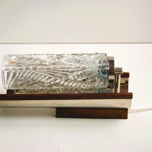K&ouml;nnte beinhalten: Eine Vintage-Wandleuchte mit einem klaren Glasschirm mit geometrischem Muster. Die Leuchte hat eine Chromoberfl&auml;che und einen Holzsockel.