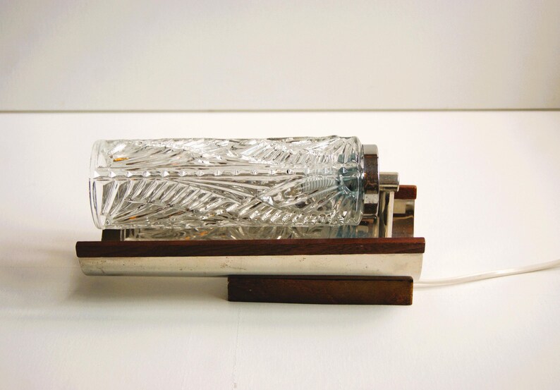 K&ouml;nnte beinhalten: Eine Vintage-Wandleuchte mit einem klaren Glasschirm mit geometrischem Muster. Die Leuchte hat eine Chromoberfl&auml;che und einen Holzsockel.