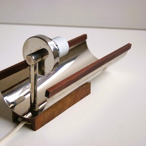 K&ouml;nnte beinhalten: Eine Wandlampe aus Silber und Holz mit einem geschwungenen Design. Die Lampe hat einen Holzsockel und einen silbernen Metallschirm. Der Schirm ist gebogen und hat einen Holzrand.
