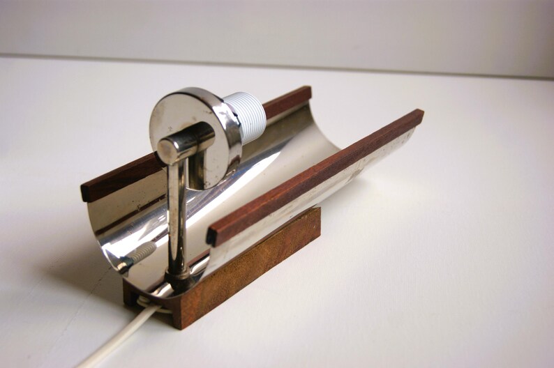 K&ouml;nnte beinhalten: Eine Wandlampe aus Silber und Holz mit einem geschwungenen Design. Die Lampe hat einen Holzsockel und einen silbernen Metallschirm. Der Schirm ist gebogen und hat einen Holzrand.