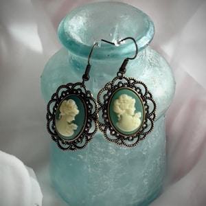 May include: A pair of antique-style earrings featuring cameo portraits. Each earring has an oval cameo with a cream-colored profile set against a teal background, framed by an ornate bronze-colored filigree. The earrings hang from bronze-colored ear wires.