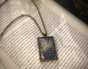 May include: A vintage-style necklace featuring a square pendant with a blue and gold peacock design. The pendant has a glass cover and is framed with a bronze-colored metal. The pendant is attached to a silver chain. The necklace is displayed on a book with the title "Pride and Prejudice" by Jane Austen.