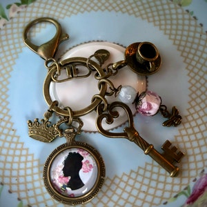 May include: Antique brass-toned keychain with various charms. Charms include a crown, a pocket watch with a silhouette, a key, a teacup, a bee, and beaded accents. The keychain is designed with a vintage aesthetic.