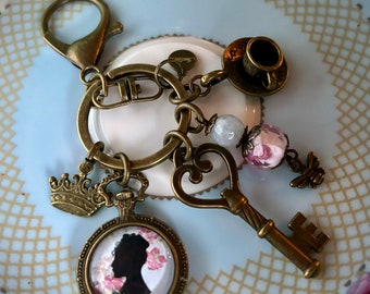 May include: Antique brass-toned keychain with various charms. Charms include a crown, a pocket watch with a silhouette, a key, a teacup, a bee, and beaded accents. The keychain is designed with a vintage aesthetic.