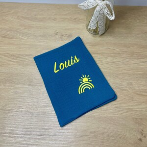 Könnte beinhalten: Eine tealblaue Musselin-Wickeldecke mit dem Namen "Louis", der in gelber Farbe gestickt ist. Ein gelbes Sonnen- und Regenbogen-Design ist ebenfalls auf die Decke gestickt.