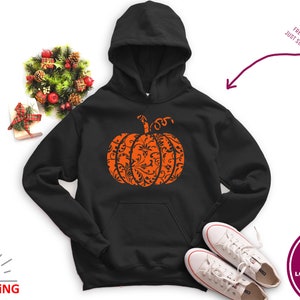 May include: Black hoodie featuring an orange pumpkin design. The pumpkin is filled with a black and white floral pattern. The hoodie has a drawstring hood and a kangaroo pocket.
