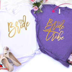 May include: Two white t-shirts with gold lettering. One shirt says "Bride" with a diamond graphic. The other shirt says "Bride Tribe".