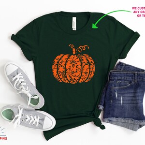 May include: A dark green t-shirt featuring an orange pumpkin design. The pumpkin is filled with a black and white floral pattern.