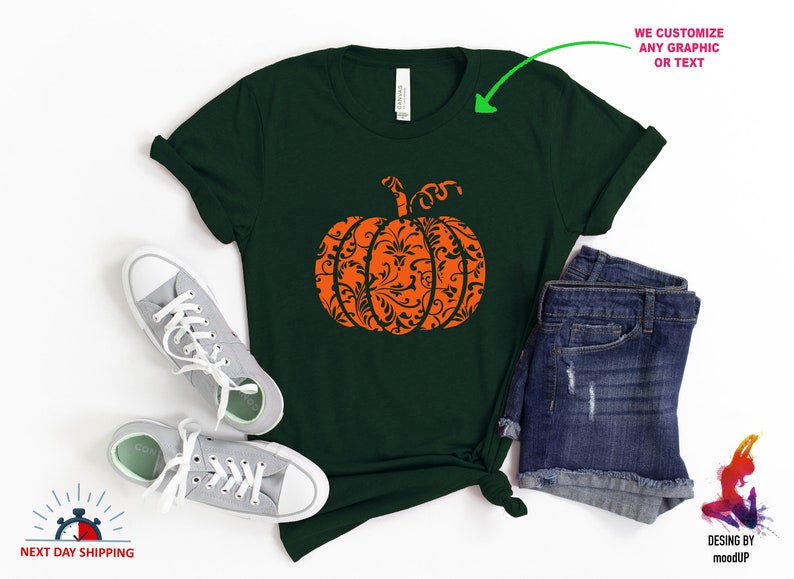 May include: A dark green t-shirt featuring an orange pumpkin design. The pumpkin is filled with a black and white floral pattern.