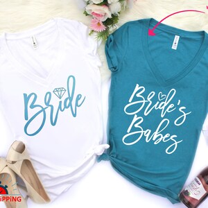 May include: Two white v-neck t-shirts with a blue and white graphic design. The shirt on the left says "Bride" with a diamond graphic. The shirt on the right says "Bride's Babes".