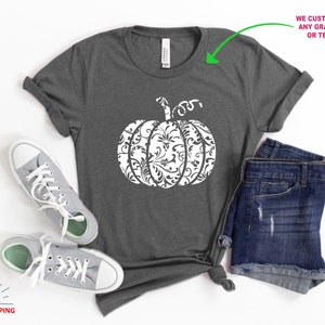 May include: A grey t-shirt featuring a white pumpkin design. The pumpkin is decorated with a white floral pattern. The shirt is displayed on a white background with a pair of grey trainers and a pair of blue denim shorts.