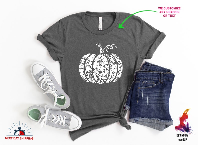 May include: A grey t-shirt featuring a white pumpkin design. The pumpkin is decorated with a white floral pattern. The shirt is displayed on a white background with a pair of grey trainers and a pair of blue denim shorts.