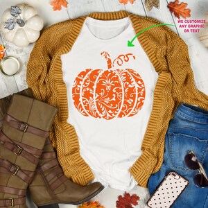 May include: White t-shirt featuring an orange pumpkin design with a white damask pattern. The pumpkin is centrally positioned on the shirt. The shirt is surrounded by autumnal items, including a brown cardigan, blue jeans, a pair of brown boots, and a white pumpkin. The text "WE CUSTOMIZE ANY GRAPHIC OR TEXT" is visible in the top right corner of the image.