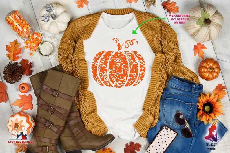 May include: White t-shirt featuring an orange pumpkin design with a white damask pattern. The pumpkin is centrally positioned on the shirt. The shirt is surrounded by autumnal items, including a brown cardigan, blue jeans, a pair of brown boots, and a white pumpkin. The text "WE CUSTOMIZE ANY GRAPHIC OR TEXT" is visible in the top right corner of the image.