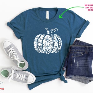 May include: A teal blue t-shirt featuring a white pumpkin design. The pumpkin is filled with a white floral pattern. The t-shirt is laid flat on a white surface with a pair of grey trainers and denim shorts.