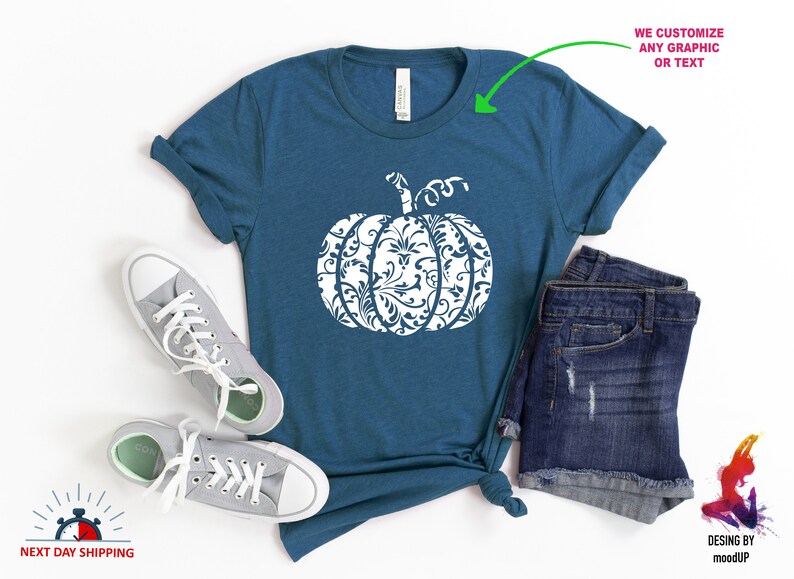 May include: A teal blue t-shirt featuring a white pumpkin design. The pumpkin is filled with a white floral pattern. The t-shirt is laid flat on a white surface with a pair of grey trainers and denim shorts.