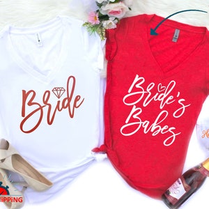 May include: Two white v-neck t-shirts with a red and white design. The shirt on the left says "Bride" with a diamond graphic. The shirt on the right says "Bride's Babes".