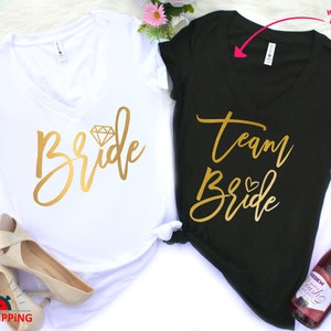 May include: Two white and black t-shirts with gold lettering. The white shirt says "Bride" with a diamond graphic. The black shirt says "Team Bride" with a small heart graphic.