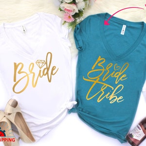 May include: Two white v-neck t-shirts with gold lettering. One shirt says "Bride" with a diamond graphic. The other shirt says "Bride Tribe".