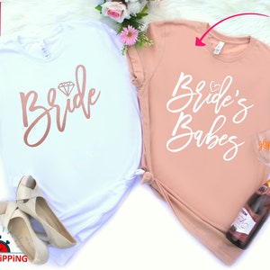 May include: Two white t-shirts with a pink and white design. The first shirt says "Bride" with a diamond graphic. The second shirt says "Bride's Babes".