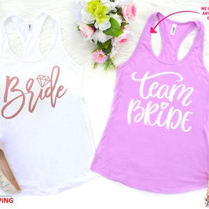 May include: Two white tank tops with the words "Bride" and "Team Bride" printed on them. The "Bride" tank top has a pink diamond graphic. The "Team Bride" tank top is pink.