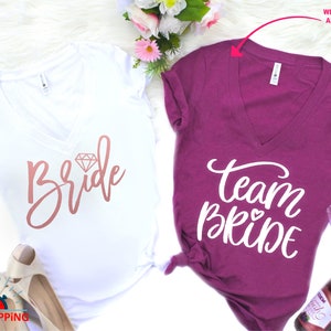 May include: Two white v-neck t-shirts with the text "Bride" and "Team Bride" in pink and white lettering. The "Bride" shirt has a diamond graphic. The "Team Bride" shirt has a small heart graphic.