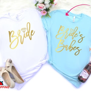 May include: Two white t-shirts with gold lettering. The first shirt says "Bride" with a diamond graphic. The second shirt says "Bride's Babes".
