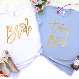 May include: Two t-shirts, one white and one light blue, with gold lettering. The white shirt says "Bride" with a diamond graphic. The light blue shirt says "Team Bride".