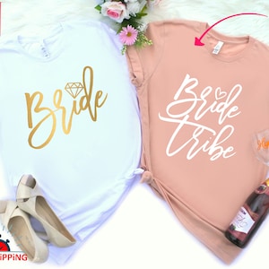 May include: Two white t-shirts with gold and white lettering. The first shirt says "Bride" with a diamond graphic. The second shirt says "Bride Tribe" with a heart graphic. The shirts are on a white fuzzy surface.