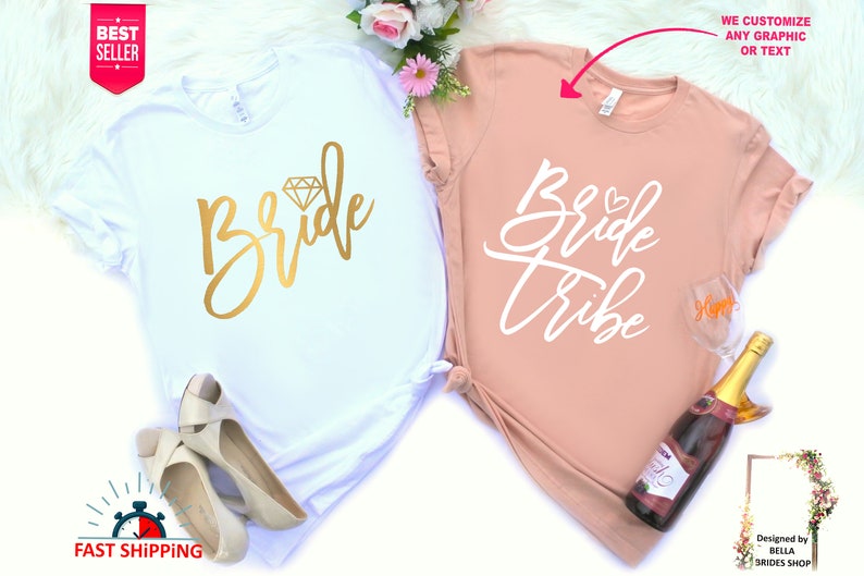 May include: Two white t-shirts with gold and white lettering. The first shirt says "Bride" with a diamond graphic. The second shirt says "Bride Tribe" with a heart graphic. The shirts are on a white fuzzy surface.