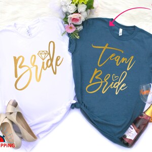May include: Two white t-shirts with gold lettering. One shirt says "Bride" with a diamond graphic. The other shirt says "Team Bride" with a heart graphic.