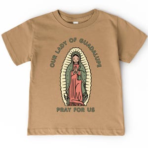 May include: Tan short-sleeve t-shirt featuring a graphic of Our Lady of Guadalupe. The illustration is in a vintage style, with the text "OUR LADY OF GUADALUPE" and "PRAY FOR US" surrounding the image.