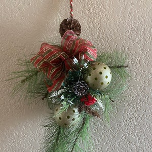 Puede incluir: Una guirnalda de pino verde con un lazo a rayas rojo y verde, adornos dorados y piñas. La guirnalda está colgada de una pared blanca.