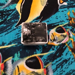 May include: A small, white square pan filled with black watercolor paint. The pan is sitting on a blue fabric with a tropical fish pattern.