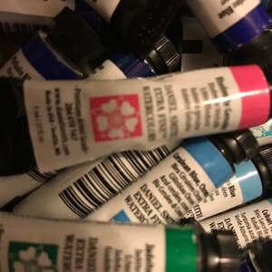 May include: A close-up of a collection of watercolor paint tubes. The tubes are various colors, including blue, green, pink, and black. The tubes are labeled with the brand name "Daniel Smith" and the words "Extra Fine Watercolors".