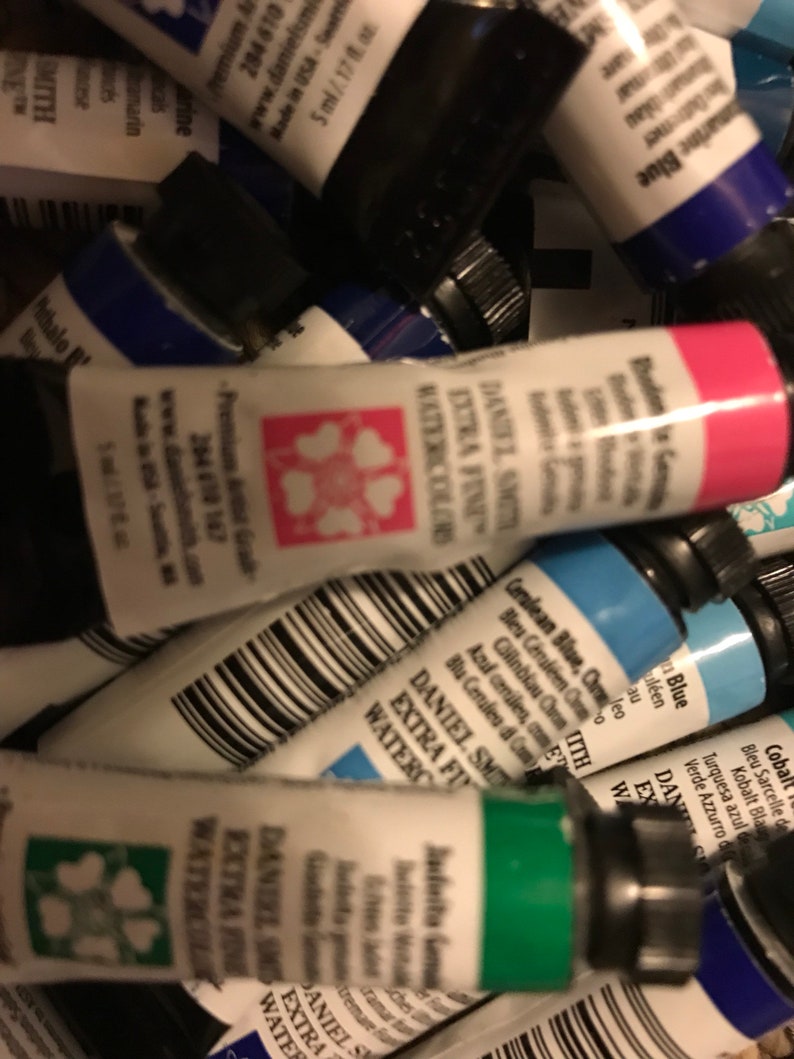 May include: A close-up of a collection of watercolor paint tubes. The tubes are various colors, including blue, green, pink, and black. The tubes are labeled with the brand name "Daniel Smith" and the words "Extra Fine Watercolors".
