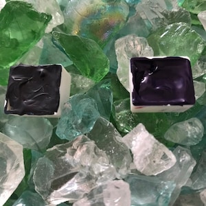 May include: Two watercolor paint pans, one black and one dark purple, are sitting on a bed of green and clear glass stones.
