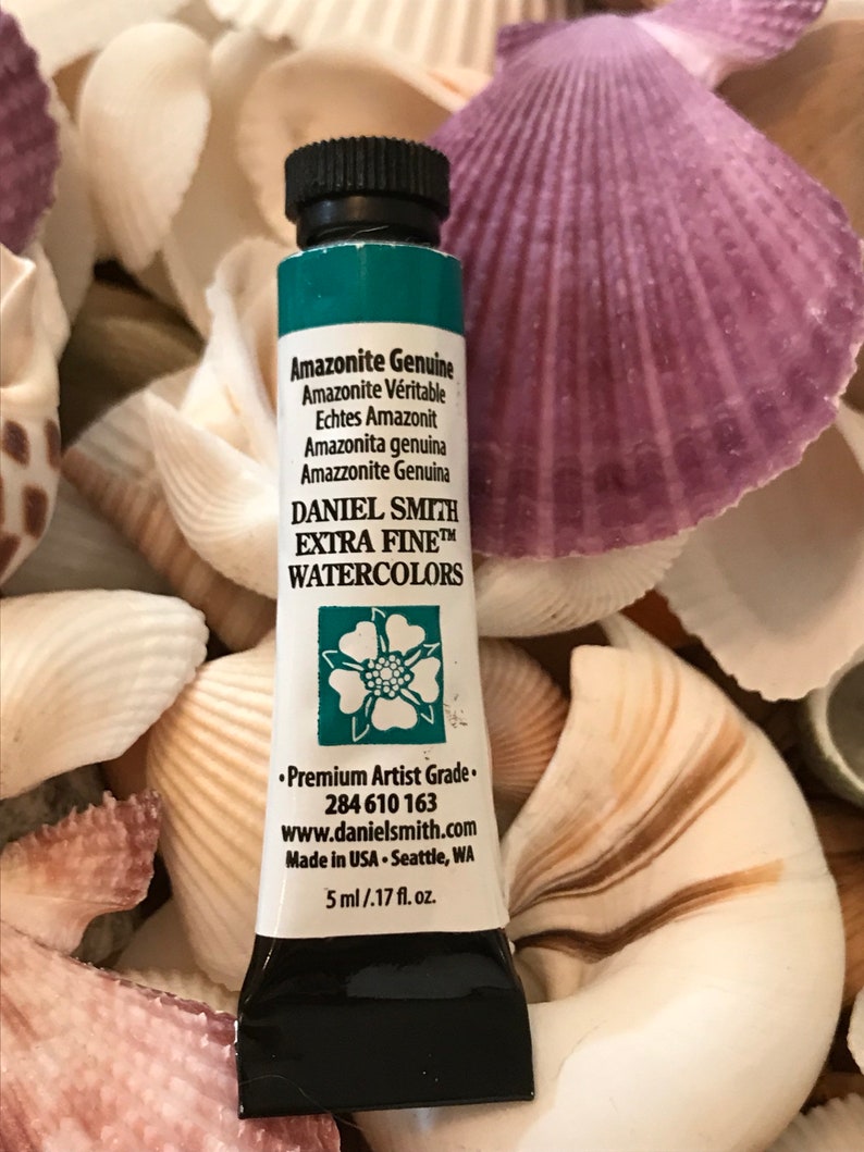 May include: A tube of Daniel Smith Extra Fine Watercolor paint in Amazonite Genuine. The tube is green with a black cap and a white label with the name of the color and the brand. The tube is sitting on a bed of seashells.