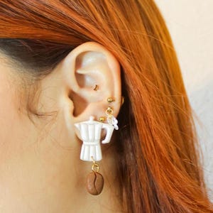 Puede incluir: Pendientes colgantes de cerámica blanca en forma de cafetera con un dije de grano de café marrón. Los pendientes cuelgan de la oreja de una persona.